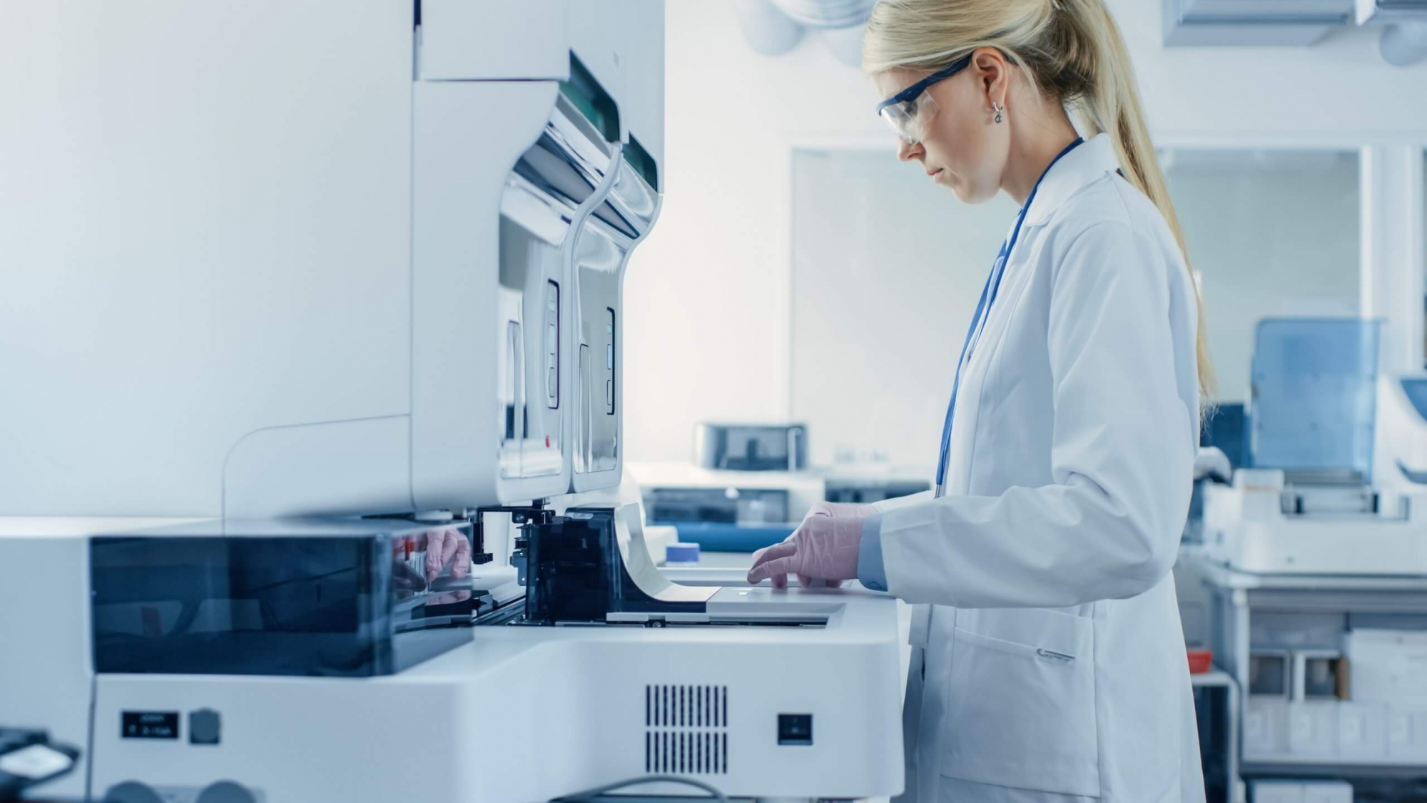 Auf dem Foto ist eine Labormitarbeiterin mit weißem Kittel und Schutzbrille zu sehen. Sie bedient ein hochmodernes Laborgerät.