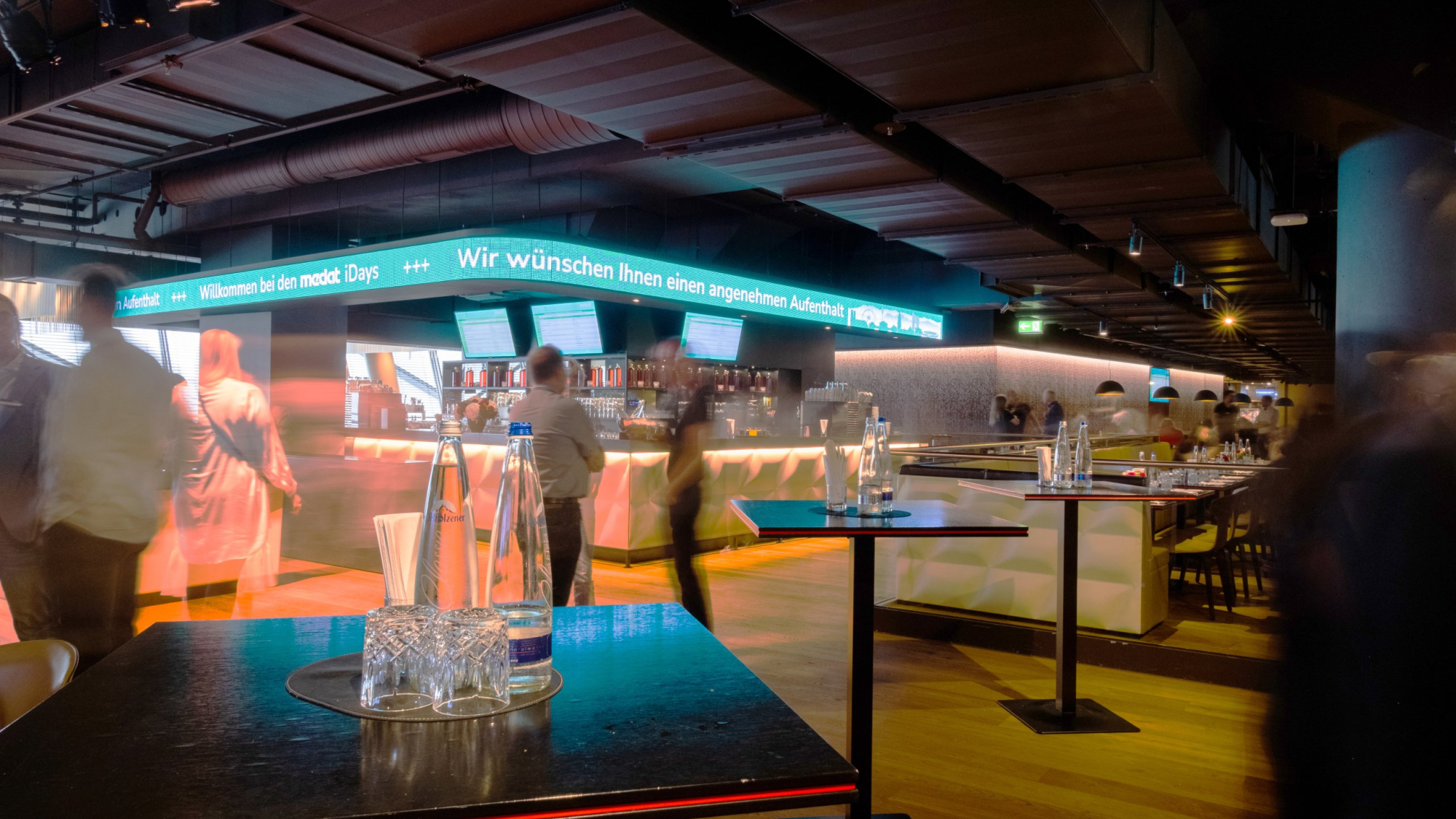 Langzeitaufnahme der Säbener Lounge in der Allianz Arena in München. Im Hintergrund die Bar und im Vordergrund 3 Stehtische mit Wasserflaschen und Gläsern.