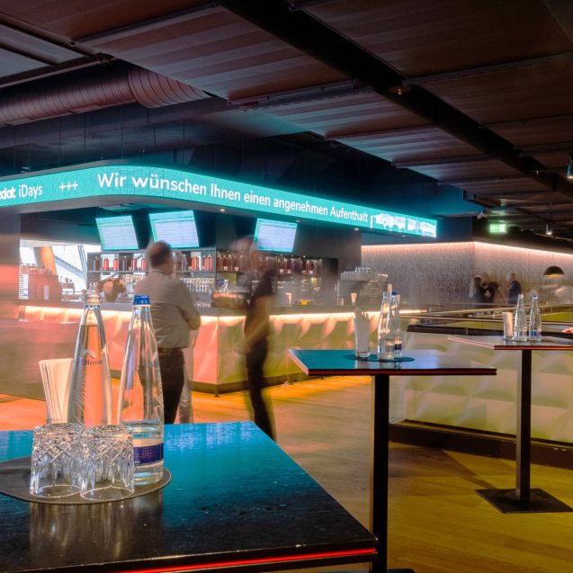 Langzeitaufnahme der Säbener Lounge in der Allianz Arena in München. ImHintergrund die Bar und im Vordergrund 3 Stehtische mit Wasserflaschen und Gläsern.